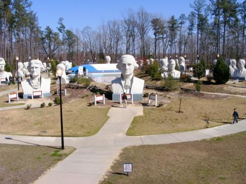 A park featuring large white busts of historical figures, surrounded by trees and pathways, with a visitor walking through.