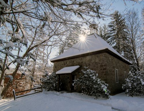 A snow-covered stone building surrounded by trees, with sunlight shining through the branches.