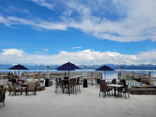 Outdoor seating area with tables and umbrellas overlooking a lake and mountains under a blue sky with clouds.