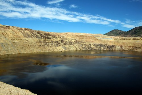 A large open-pit mine with steep, layered walls and a calm water-filled bottom under a blue sky.