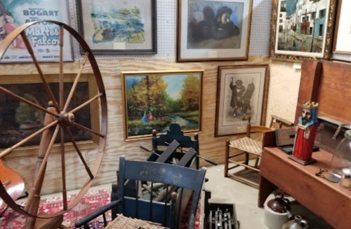 A cozy antique shop filled with vintage furniture, paintings, and a large wooden wheel in a warm, inviting setting.