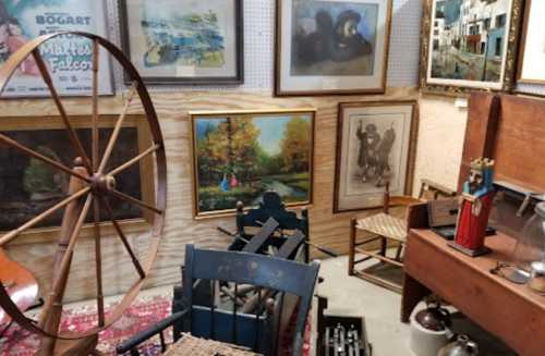 A cozy antique shop filled with vintage furniture, paintings, and a large wooden wheel in a warm, inviting setting.