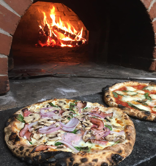 Two pizzas in front of a brick oven, one topped with ham, mushrooms, and onions, the other with tomato and cheese.