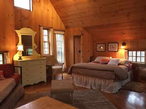 Cozy wooden cabin interior featuring a bed, dresser, and seating area with warm lighting and large windows.