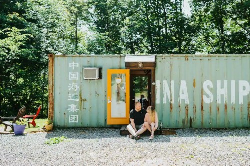 A couple sits outside a rustic shipping container in a wooded area, enjoying a sunny day.