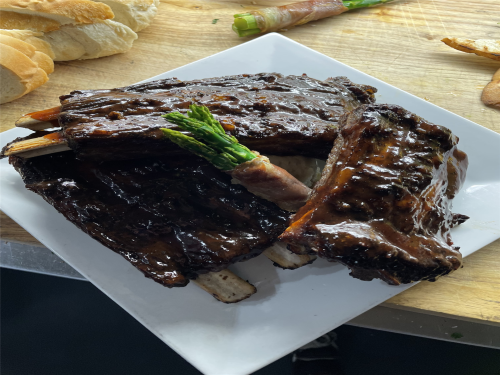 A plate of glazed ribs garnished with an asparagus spear, served alongside slices of bread.