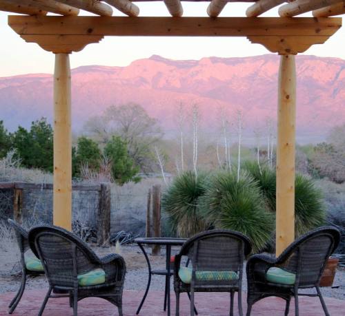 A wooden pergola frames a view of mountains at sunset, with green plants and wicker chairs in the foreground.