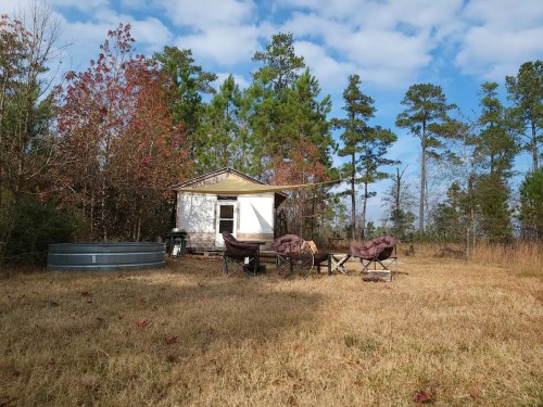 A small cabin surrounded by trees, with outdoor seating and a water tank in a grassy area under a partly cloudy sky.