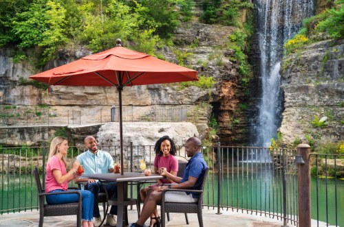 Four friends enjoy drinks at a table under an umbrella, with a waterfall and lush greenery in the background.