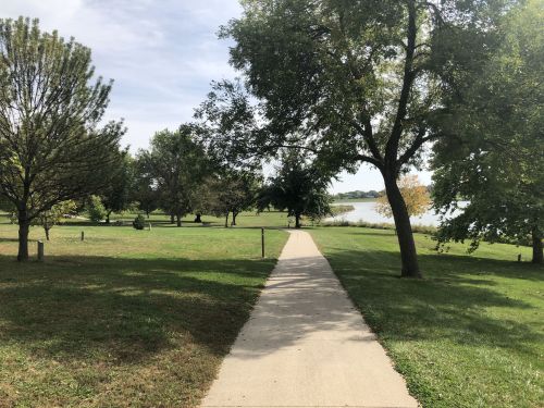 A paved path leads through a grassy park with trees, ending near a calm lake under a partly cloudy sky.