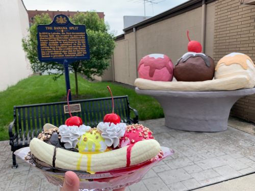 A hand holds a banana split topped with cherries, with a large banana split sculpture and historical sign in the background.