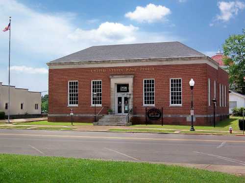 Historic brick building with large windows, labeled "United States Post Office," set against a blue sky.