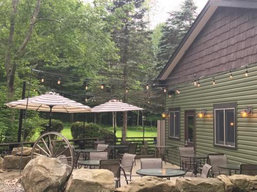 A cozy outdoor patio with umbrellas, string lights, and a rustic wheel, surrounded by trees and a green building.