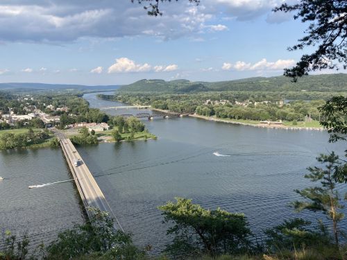 A scenic view of a river with bridges, lush green hills, and a small town in the background under a partly cloudy sky.