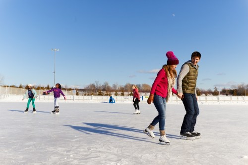 A couple skates hand-in-hand on an outdoor rink, surrounded by others enjoying the winter day.