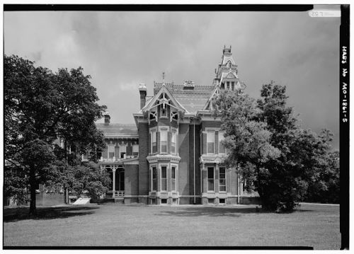 Historic brick mansion with intricate architectural details, surrounded by trees and a grassy lawn under a cloudy sky.