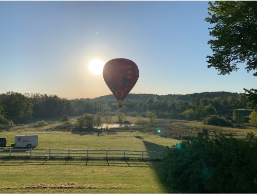 A colorful hot air balloon floats above a serene landscape at sunrise, with a pond and trees in the background.