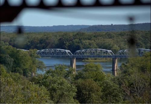 A view of a metal bridge spanning a river, surrounded by lush green trees and distant hills under a clear sky.