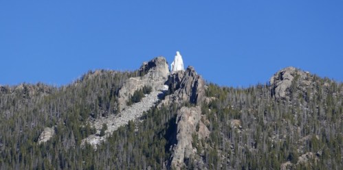 A white statue of a figure stands atop a rocky mountain, surrounded by evergreen trees and a clear blue sky.