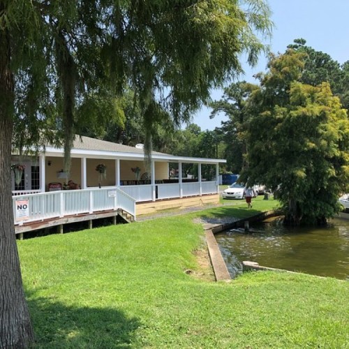 A waterfront house with a porch, surrounded by greenery, and a small dock leading to the water.