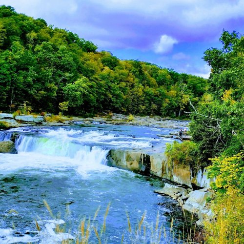 A serene river flows over rocks, surrounded by lush green trees and a cloudy blue sky.