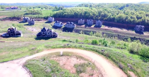 Aerial view of abandoned, partially constructed houses in a green landscape with rolling hills in the background.