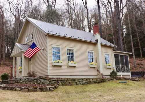 A charming beige house with a metal roof, American flag, and flower boxes, surrounded by trees and a grassy yard.