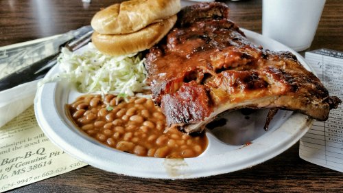 A plate of barbecue ribs with baked beans, coleslaw, and two sandwich buns on a paper plate.