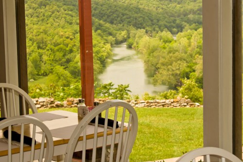 A serene view of a river surrounded by lush greenery, seen through a window with white chairs in the foreground.