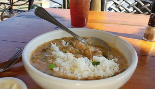A bowl of gumbo with rice, a spoon, and a drink in the background on a wooden table.