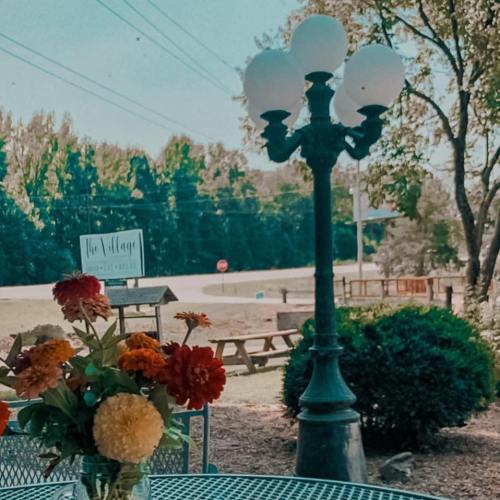A table with a flower vase in front of a lamp post, surrounded by greenery and a sign for "The Village."