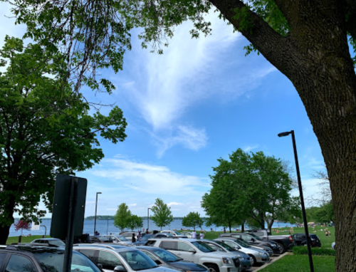 A scenic view of a lake with trees and parked cars under a blue sky with clouds.