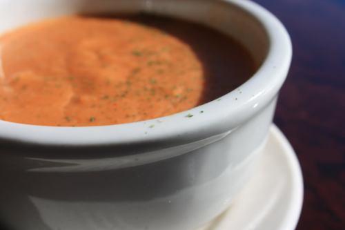 A close-up of a bowl of creamy tomato soup, garnished with herbs, on a saucer against a wooden table background.