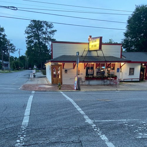 A small, charming restaurant at a street corner, with outdoor seating and trees in the background, during twilight.