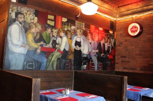 A cozy restaurant interior featuring a mural of people at a bar, with wooden booths and red table settings.