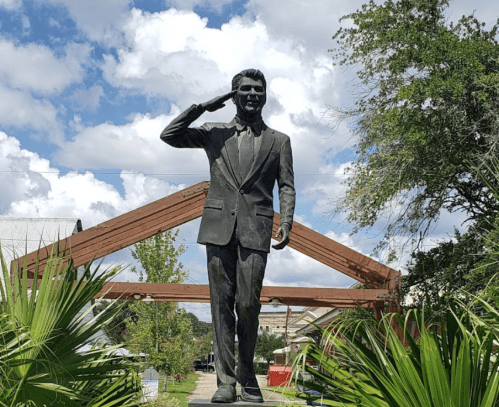 A bronze statue of a man in a suit saluting, set against a backdrop of blue skies and greenery.