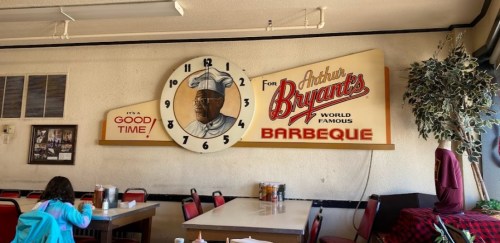 Interior of a barbecue restaurant featuring a large clock and a sign for Arthur Bryant's Barbeque. Red chairs and tables visible.