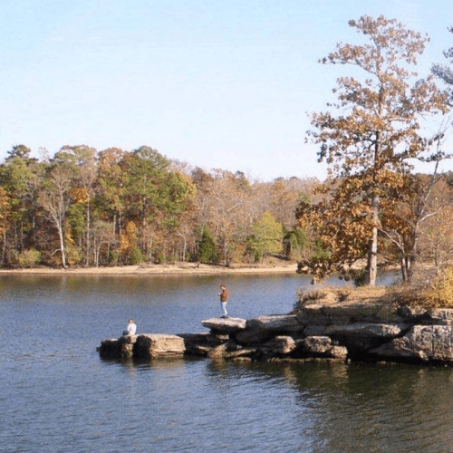 A person stands on a rocky shore by a calm lake, surrounded by trees with autumn foliage.