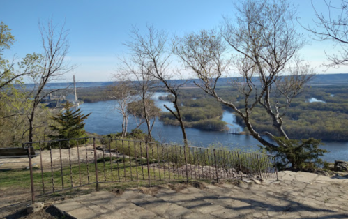 A scenic view of a river surrounded by trees, with a clear blue sky and a railing in the foreground.