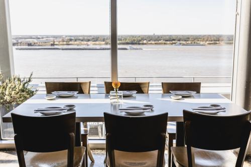 A dining table set for a meal, overlooking a river view through large windows.