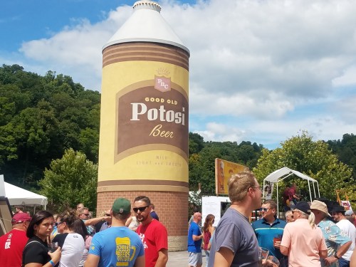 A large Potosi Beer tower stands at a festival, with people enjoying the event under a blue sky.