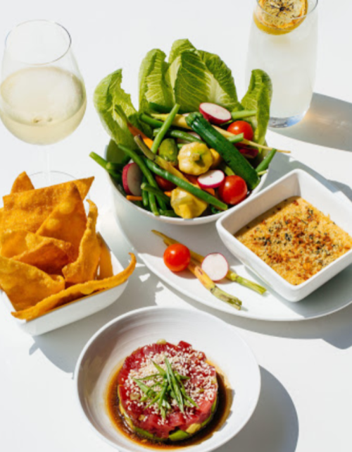 A vibrant meal featuring a salad, tuna tartare, baked dish, and crispy chips, accompanied by a glass of white wine.