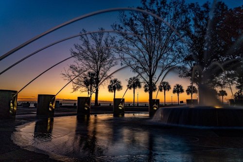 A serene sunset scene with a fountain, silhouetted trees, and palm trees against a colorful sky.