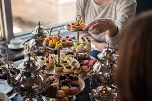 A tiered stand filled with colorful pastries and desserts, with a person reaching for a treat. Silver teapots are nearby.