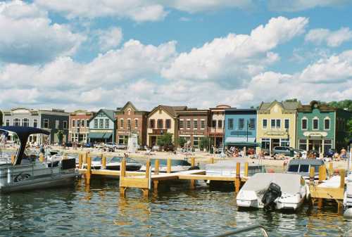 Colorful waterfront buildings line a marina, with boats docked and people enjoying the beach under a partly cloudy sky.