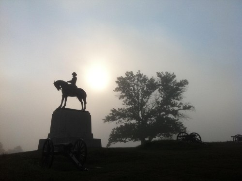 Silhouette of a horse statue with a rider against a foggy background, sun peeking through, and cannons nearby.