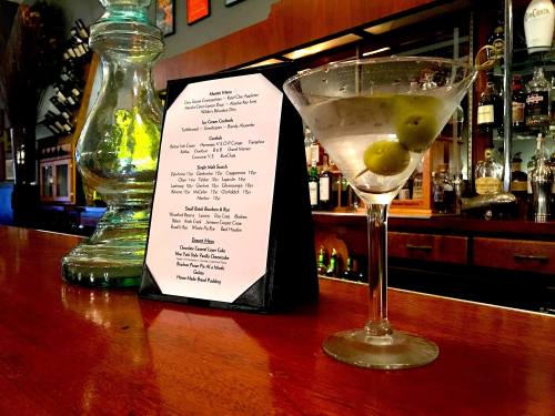 A martini with olives sits on a bar counter next to a menu, with a vintage lamp in the background.