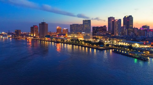 Aerial view of a city skyline at dusk, with buildings reflecting on a river and lights illuminating the waterfront.