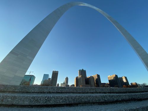 A wide-angle view of the Gateway Arch with a city skyline in the background under a clear blue sky.