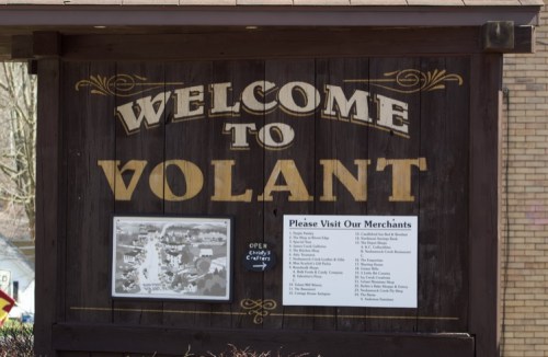 Wooden sign reading "Welcome to Volant" with a list of local merchants and an open hours notice.